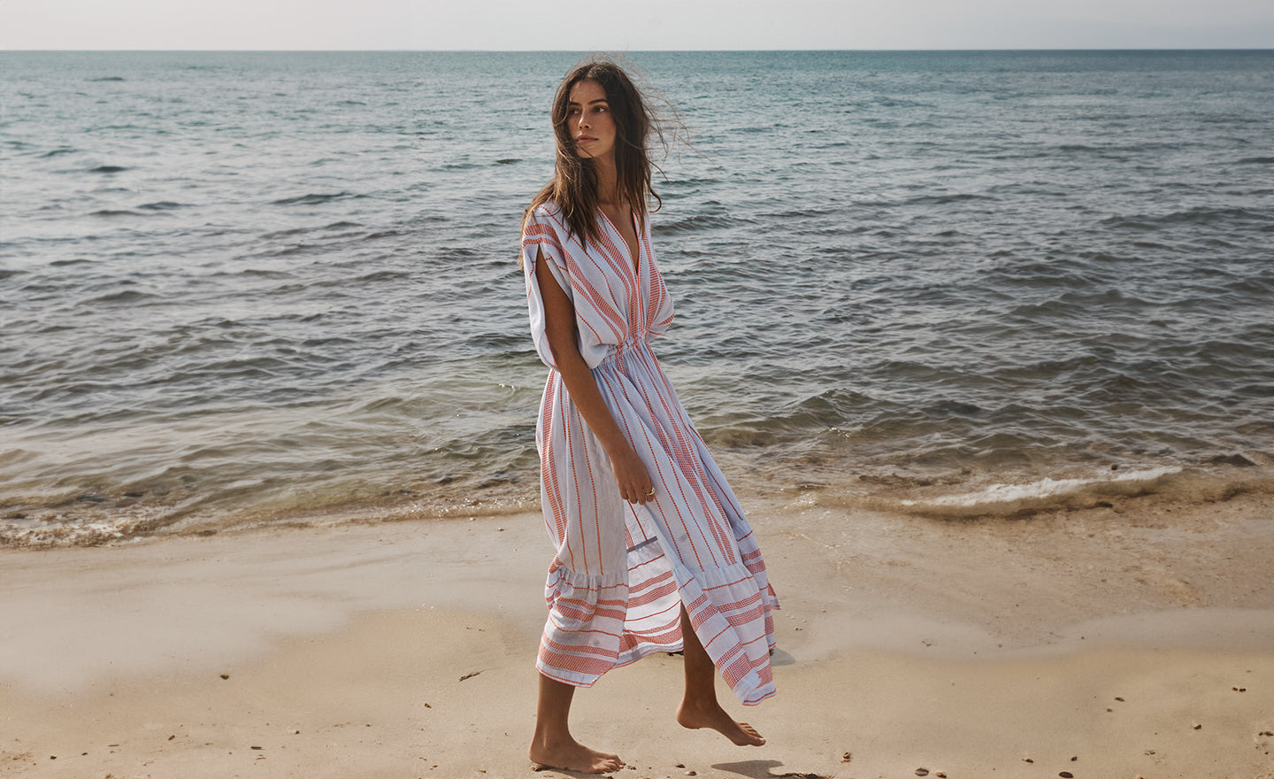 Woman Walking on the beach wearing lemlem plunge dress featruring dotted stripe pattern in orange color on pale blue background