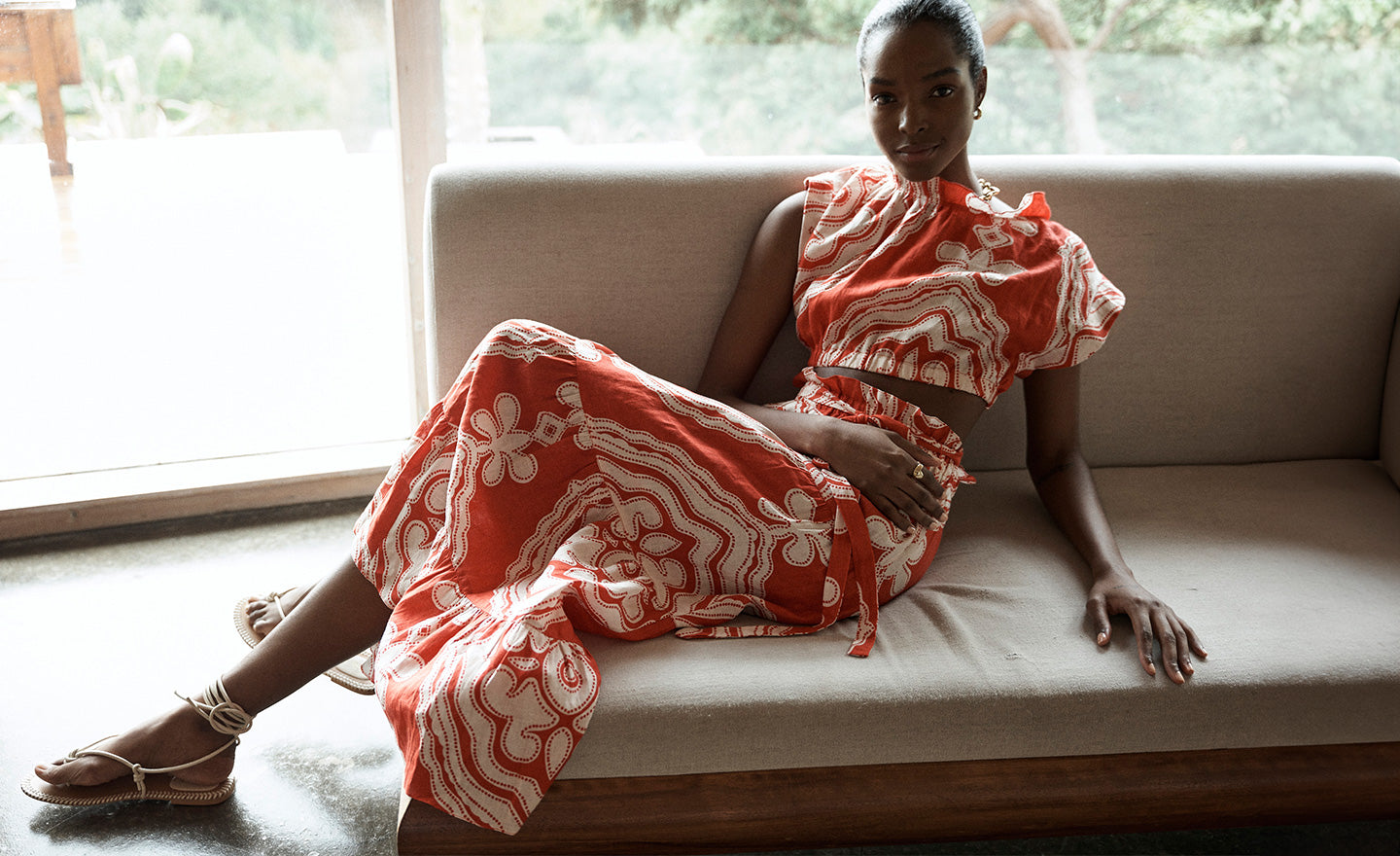 Woman Sitting on the couch wearing lemlem cropped top and maxi skirt featuring ancient inspired pattern in white color on red background