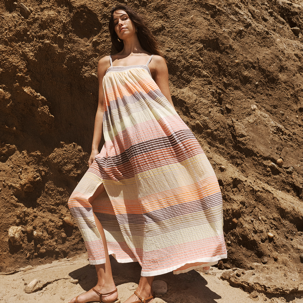 Woman standing a rock wearing a maxi lemlem dress with thine gradual brown to yellow stripes.