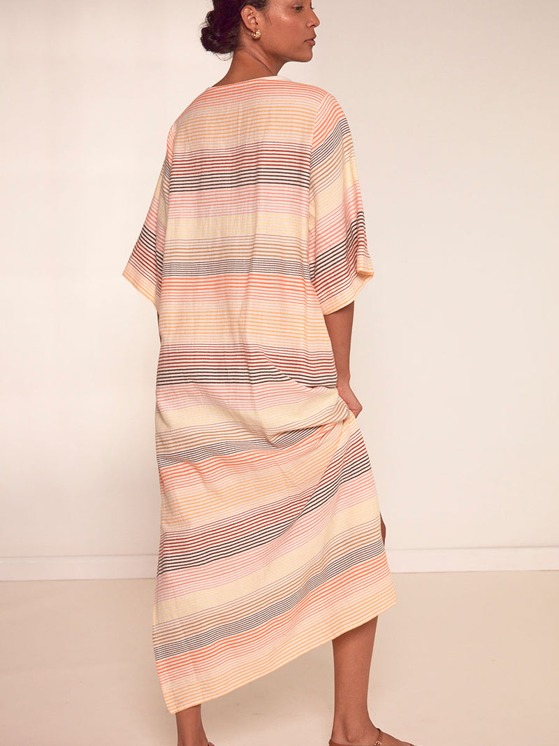 Back View of a Woman Standing Wearing lemlem Edna Dress Featuring continuous stripe pattern in warm yellow, orange and peach tones.