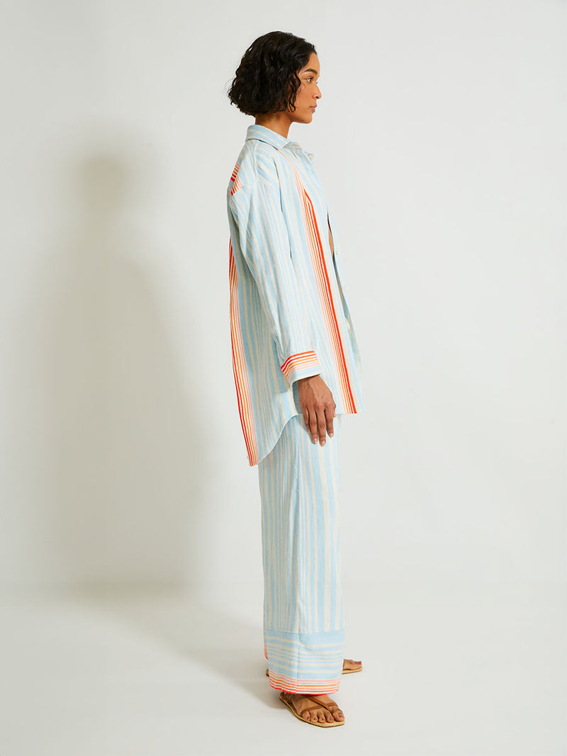 Side of a Woman Standing Wearing lemlem Desta Pants and Mariam Shirt featuring soft, variegated two-color stripe in sky blue and cream, complemented by placed bands of bright orange and tangerine