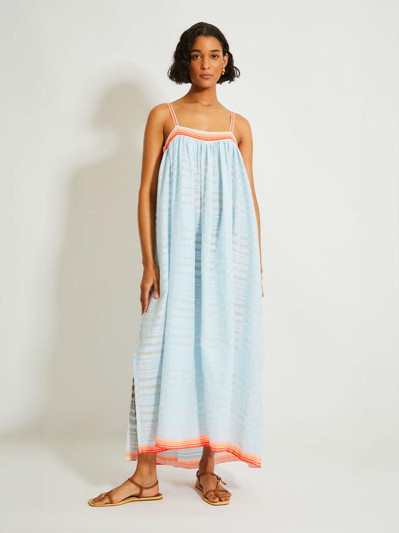 Woman Standing Wearing lemlem EDA Slip Dress featuring soft, variegated two-color stripe in sky blue and cream, complemented by placed bands of bright orange and tangerine.