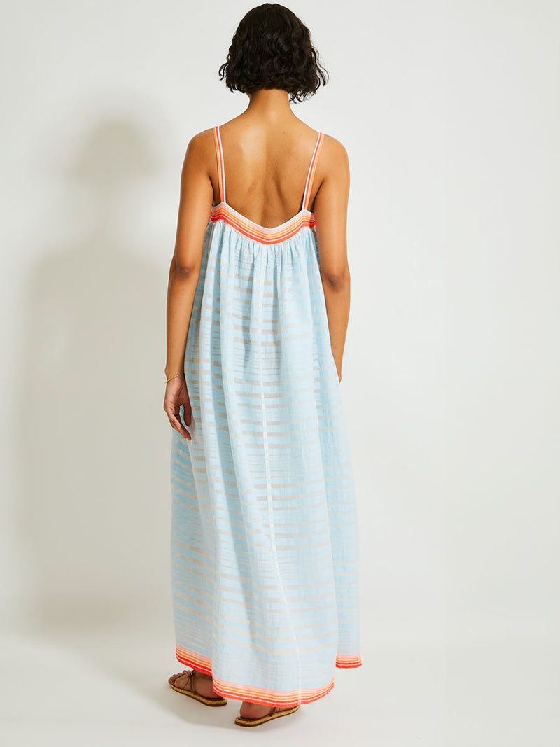 Back of a Woman Standing Wearing lemlem EDA Slip Dress featuring soft, variegated two-color stripe in sky blue and cream, complemented by placed bands of bright orange and tangerine.