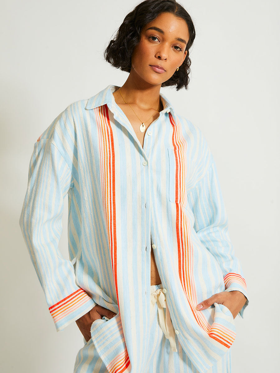 Woman Standing Wearing lemlem Mariam Shirt Featuring soft, variegated two-color stripe in sky blue and cream, complemented by placed bands of bright orange and tangerine.