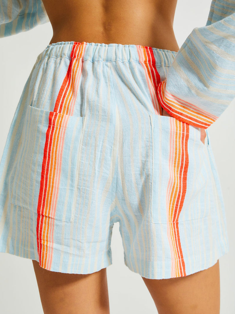 Close up on Woman's Back Standing Wearing lemlem SAFIA Shorts featuring soft, variegated two-color stripe in sky blue and cream, complemented by placed bands of bright orange and tangerine