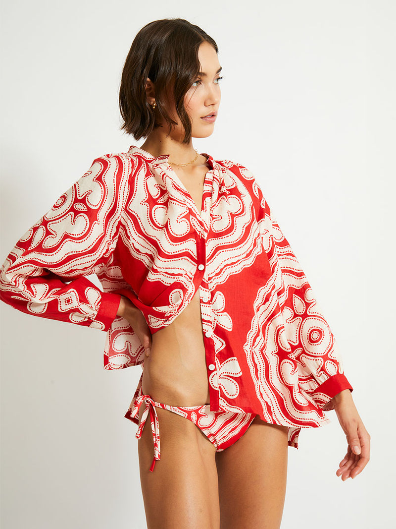 Side View of a Woman Standing Wearing lemlem MITA Blouse featuring custom-designed print inspired by ancient architectural patterns in rich, vibrant red and creamy ivory color and matching string bottom