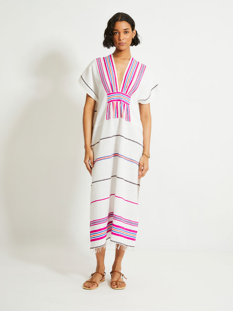 Woman Standing Wearing lemlem Gasira Caftan featuring stripe pattern of bright magenta, blue, and orange on s crispy white background.