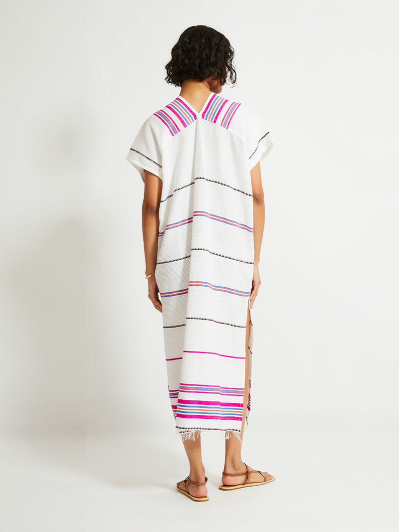 Back of a Woman Standing Wearing lemlem Gasira Caftan featuring stripe pattern of bright magenta, blue, and orange on s crispy white background.