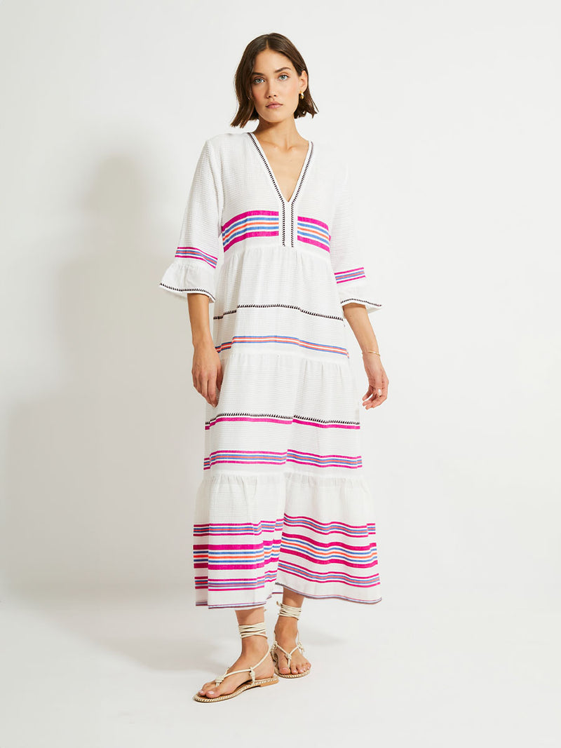 Woman Standing Wearing lemlem HAWI Flutter Dress featuring stripe pattern of bright magenta, blue, and orange on s crispy white background.