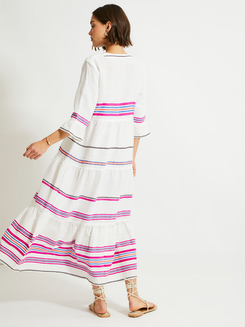 Back of a Woman Standing Wearing lemlem HAWI Flutter Dress featuring stripe pattern of bright magenta, blue, and orange on s crispy white background.