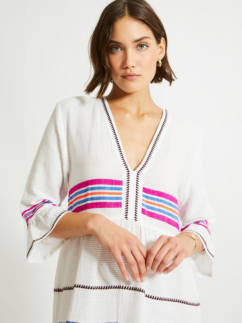 Woman Standing Wearing lemlem HAWI Flutter Dress featuring stripe pattern of bright magenta, blue, and orange on s crispy white background.