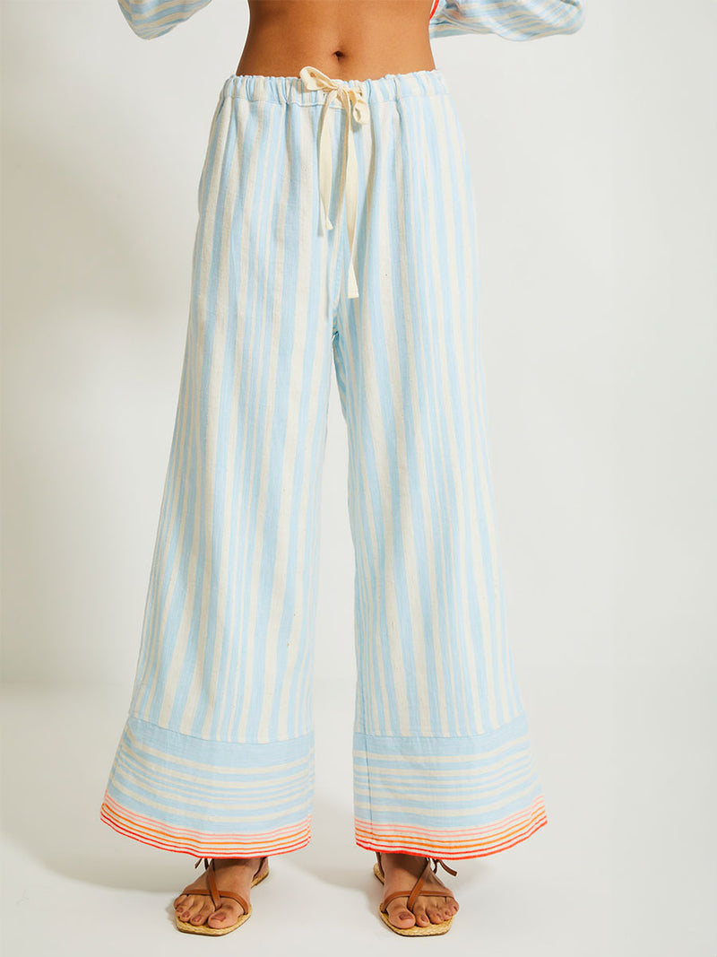 Close up on a Woman Standing Wearing lemlem Desta Pants and Mariam Shirt featuring soft, variegated two-color stripe in sky blue and cream, complemented by placed bands of bright orange and tangerine