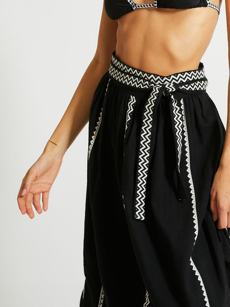 Close up on a Woman Standing Wearing lemlem TOLA Skirt Featuring rich black background with intricate cream Tibeb border pattern and black triangle bikini top