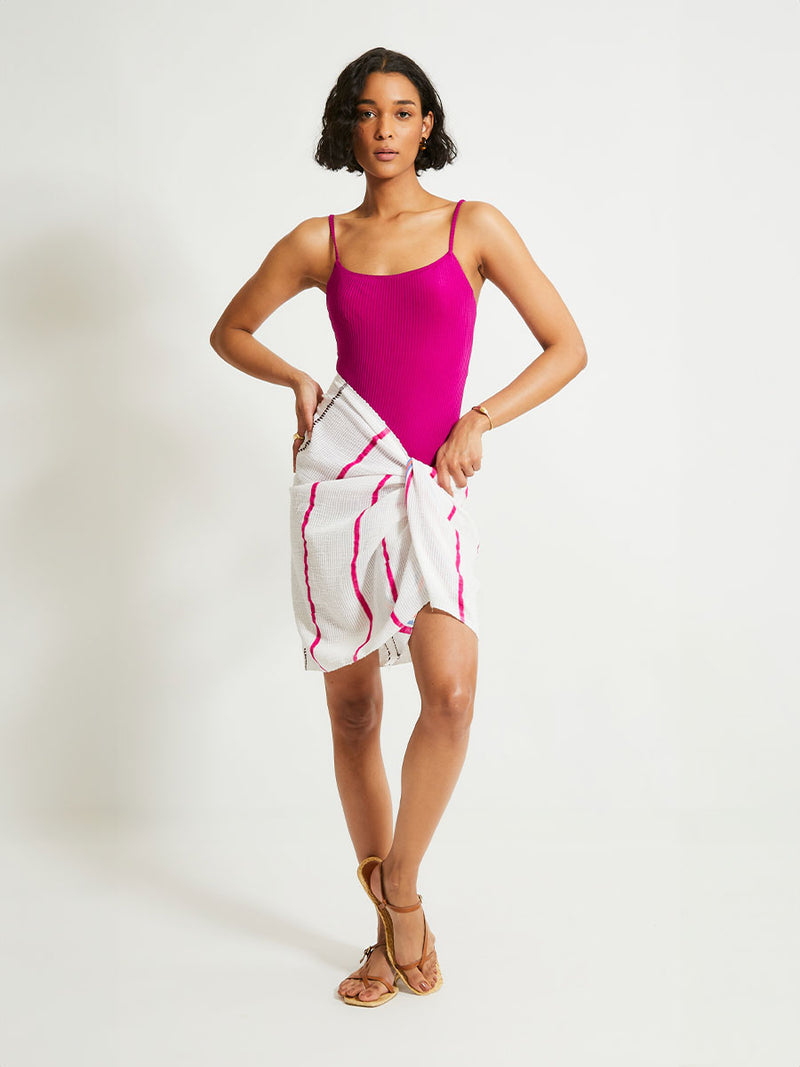 Woman Standing Wearing lemlem LEMA Sarong featuring stripe pattern of bright magenta, blue, and orange on s crispy white background and ELENE One Piece Swumsuit featuring magenta color