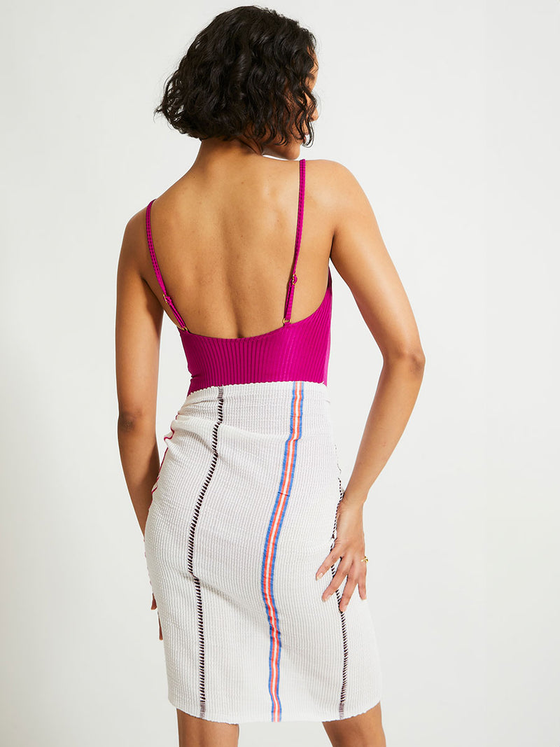 Back of a Woman Standing Wearing lemlem LEMA Sarong featuring stripe pattern of bright magenta, blue, and orange on s crispy white background and ELENE One Piece Swumsuit featuring magenta color