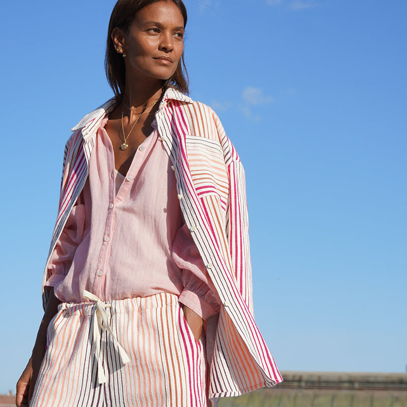 Liya Kebede standing on a New York city roof wearing the new lemlem Mariam shirt with a light pink mita blouse under and matching pants in white with thin pink and orange stripes.