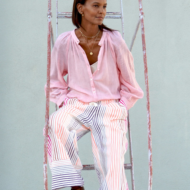 Liya Kebede standing sitting on a ladder wearing the new lemlem Mita blouse and lemlem Desta pants in white with thin pink and orange stripes.