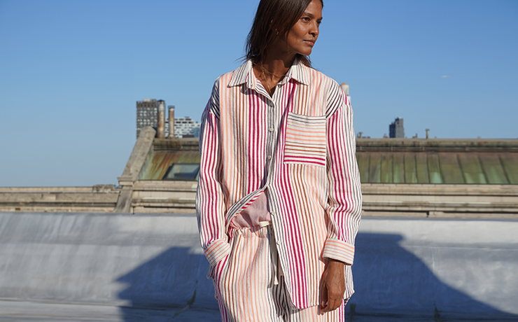 Liya Kebede standing in front on a New York city roof wearing the new lemlem Mariam shirt and matching pants in white with thin pink and orange stripes.