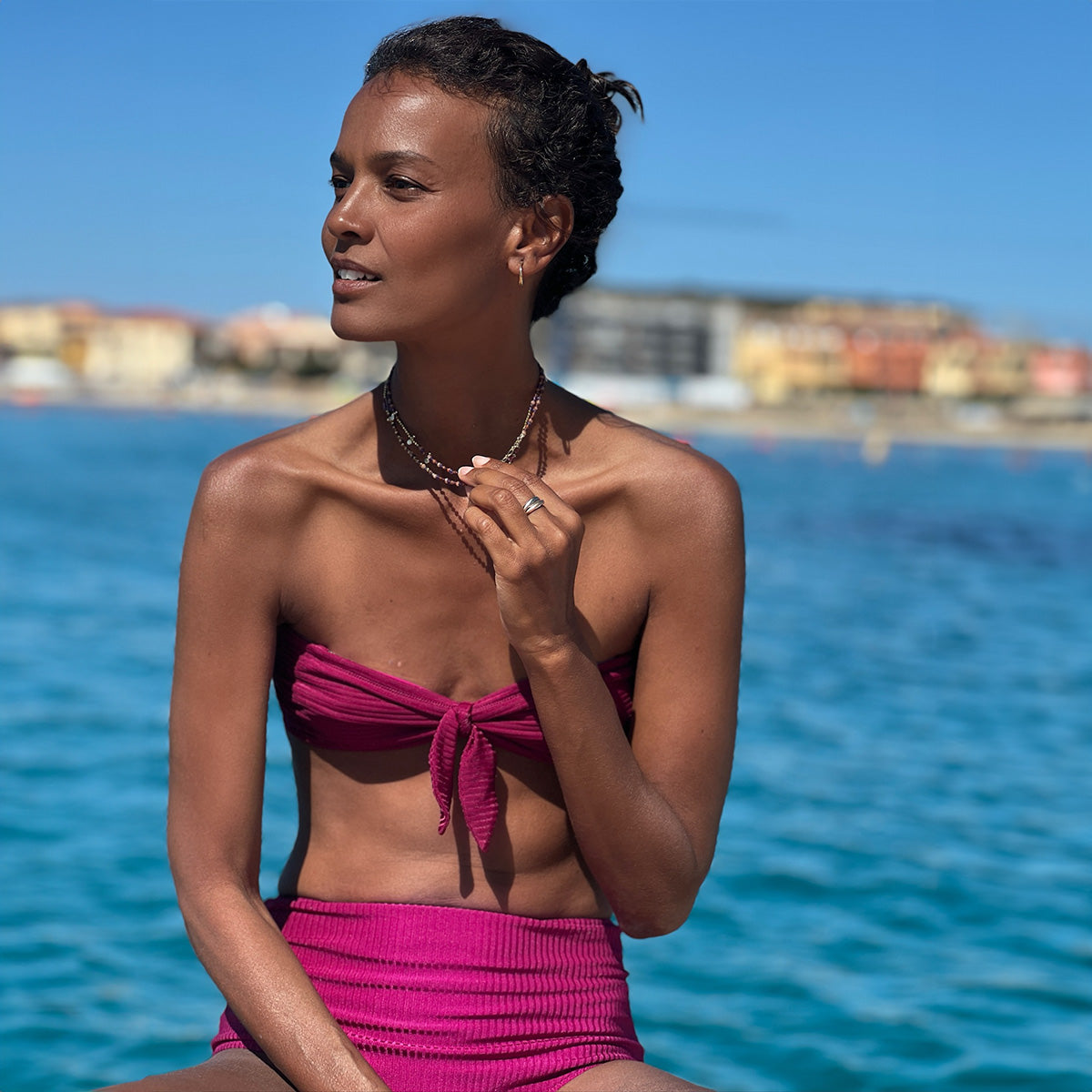 Liya sitting wearing lemlem beri magenta bandeau top and high waist bottom featuring ribbed fabric in bright berry magenta color