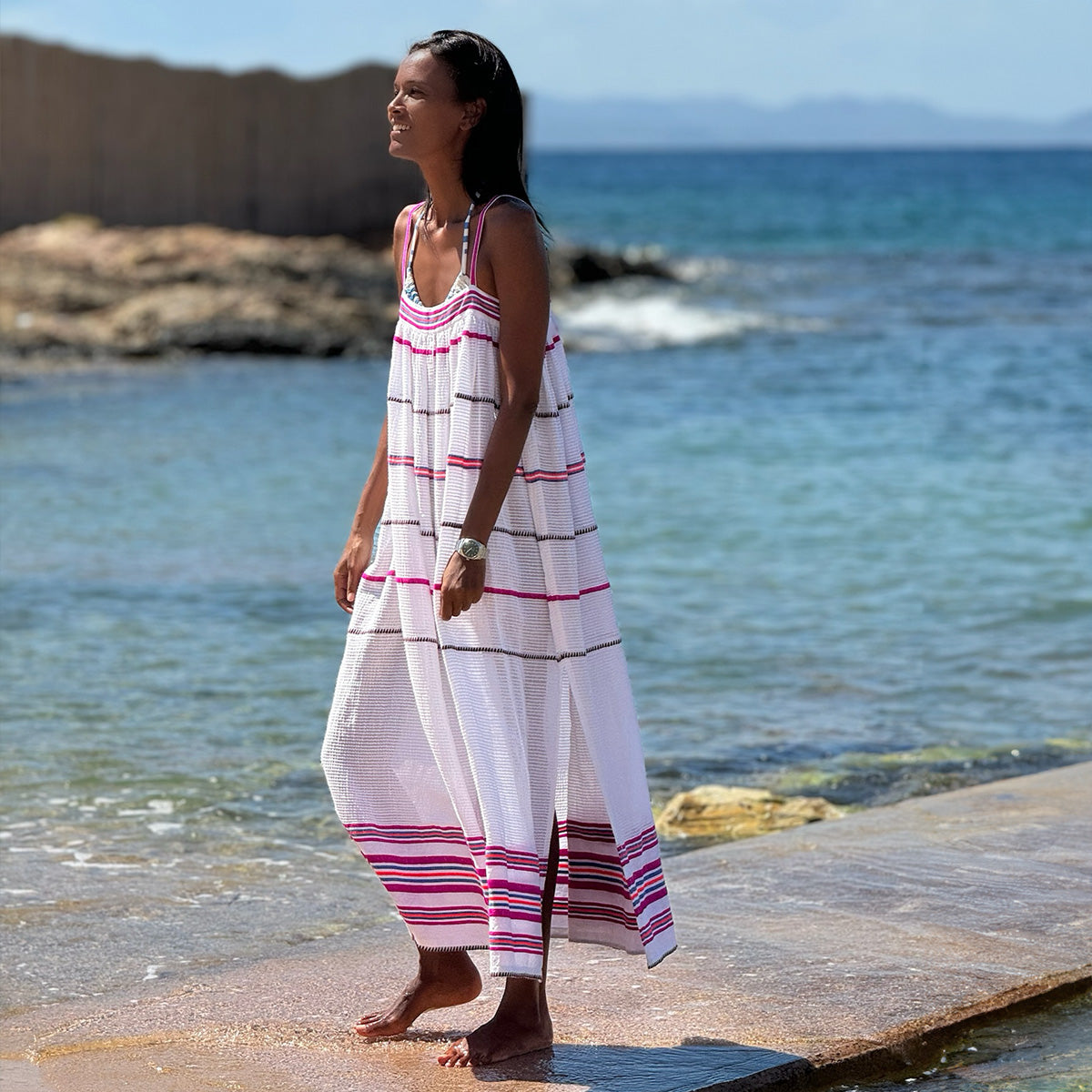 Liya walking by the sea wearing lemlem Eda Flowy Slip Dress featuring stripe pattern in orchid, black, blue and orange colors on white crispy background