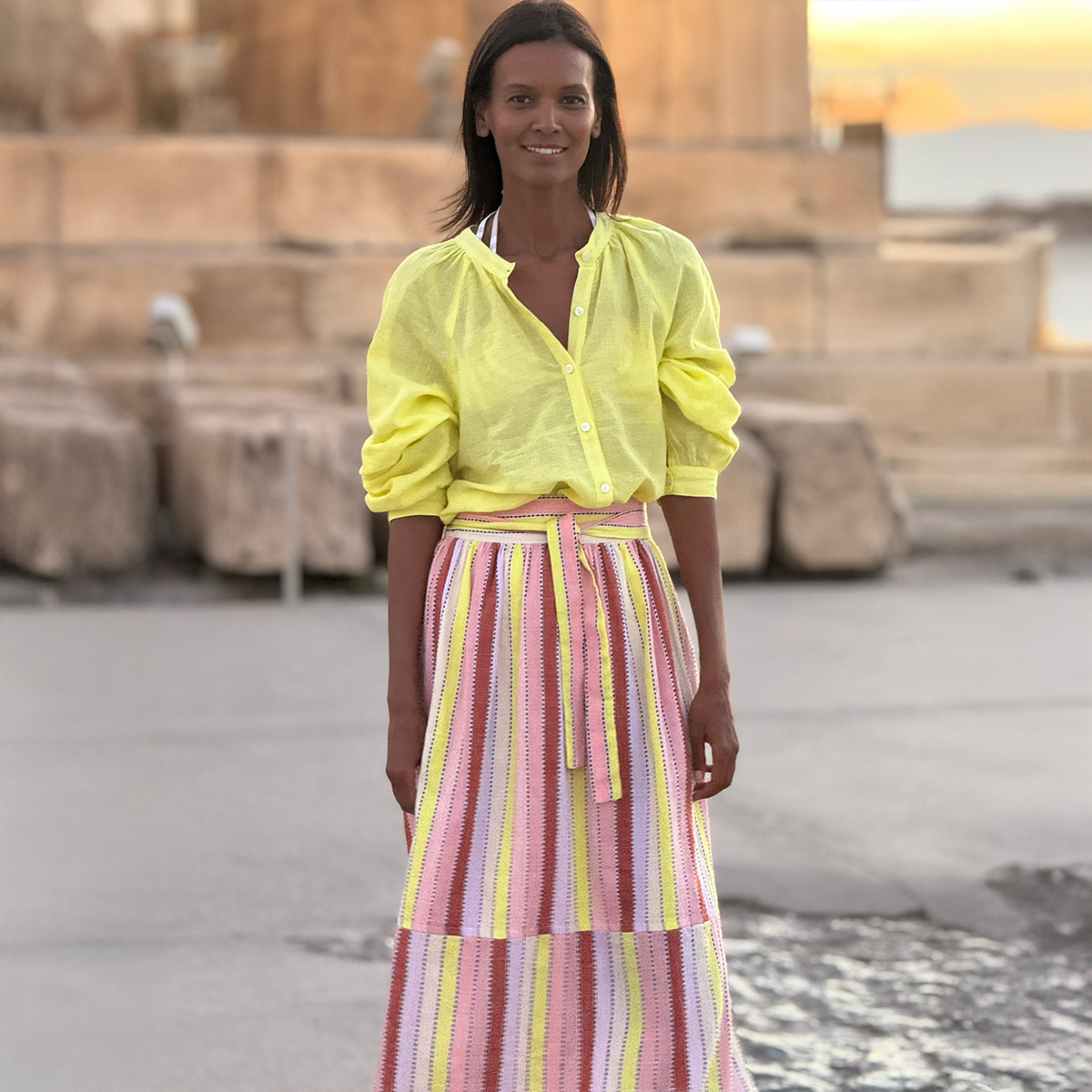 Liya Kebede standing in front of greek ruins wearing a lemlem hand woven blouse in yellow and a lemlem hand woven skirt with pink, white, yellow and terra cotta vertical stripes.