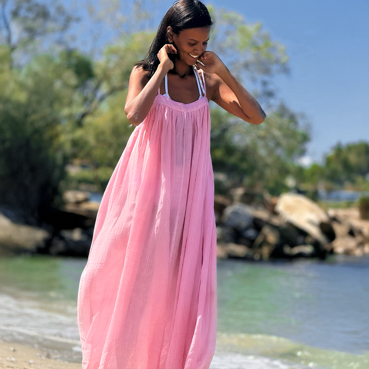 Liya Kebede smiling while walking on a beach putting her hair back behind her hears and wearing a lemlem hand woven maxi slip dress in light pink.