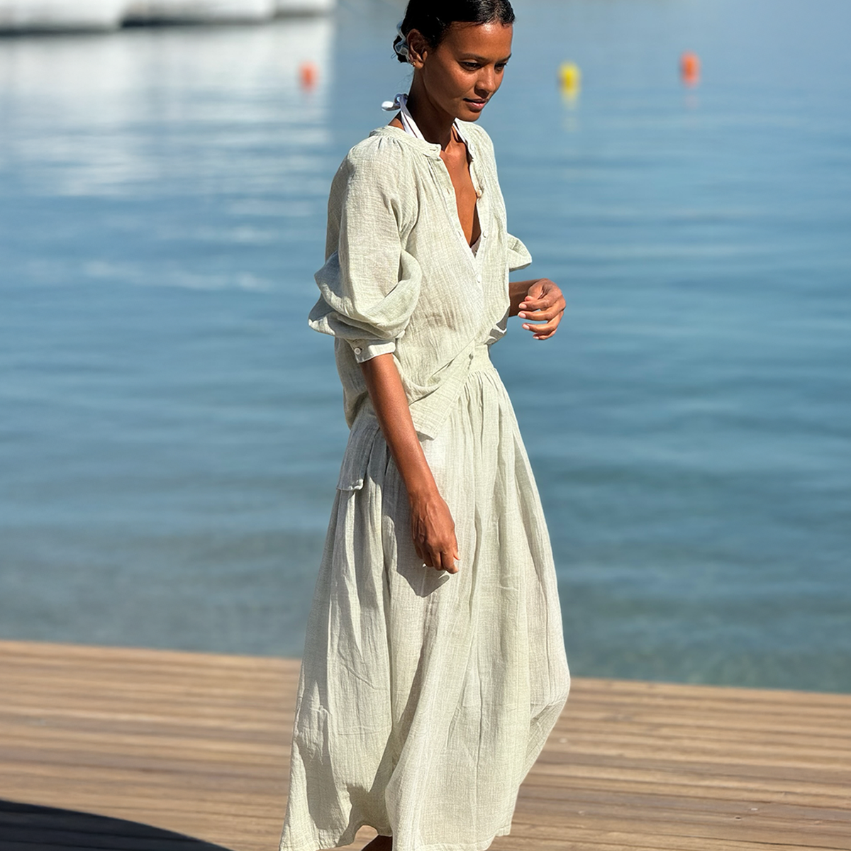 Liya Kebede walking on a deck with the sea in the background wearing a lemlem hand woven blouse in sage and matching hand woven skirt. 