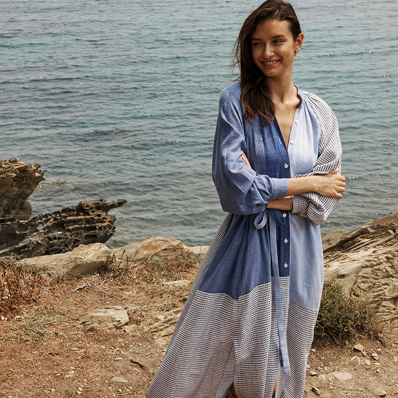 Woman standing a cliff with the Mediterranean sea in the background wearing a long lemlem handwoven dress in blue.