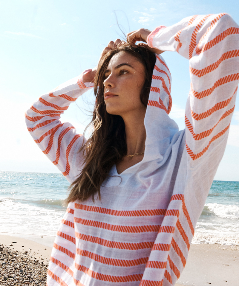 Woman standing on a beach in Montauk, NY wearing a light blue with graphic red stripes lemlem hoodie.