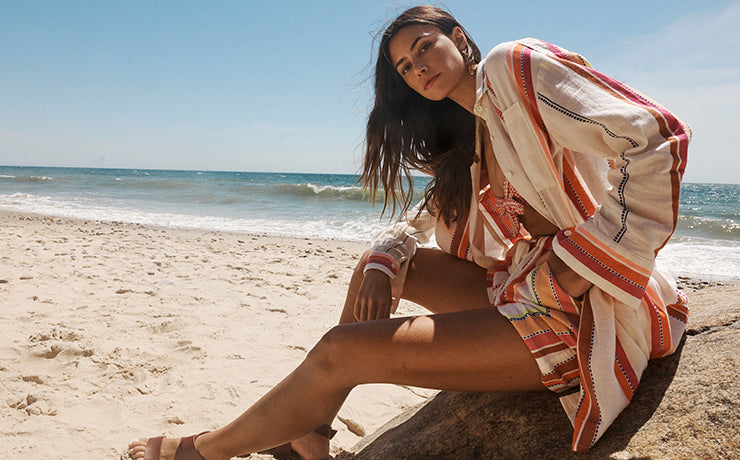 Woman sitting on a rock on a beach in Montauk, NY wearing a pink lemlem bandeau bikini top under an open lemlem shirt and matching lemlem short in cream with pink and orange graphic stripes.