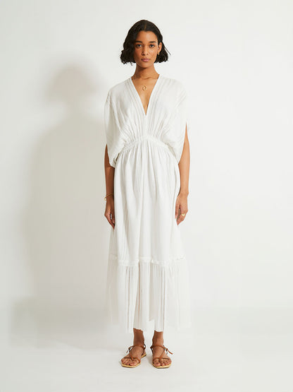 Woman standing wearing a white Abira plunge neck maxi dress with stitches of silver lurex