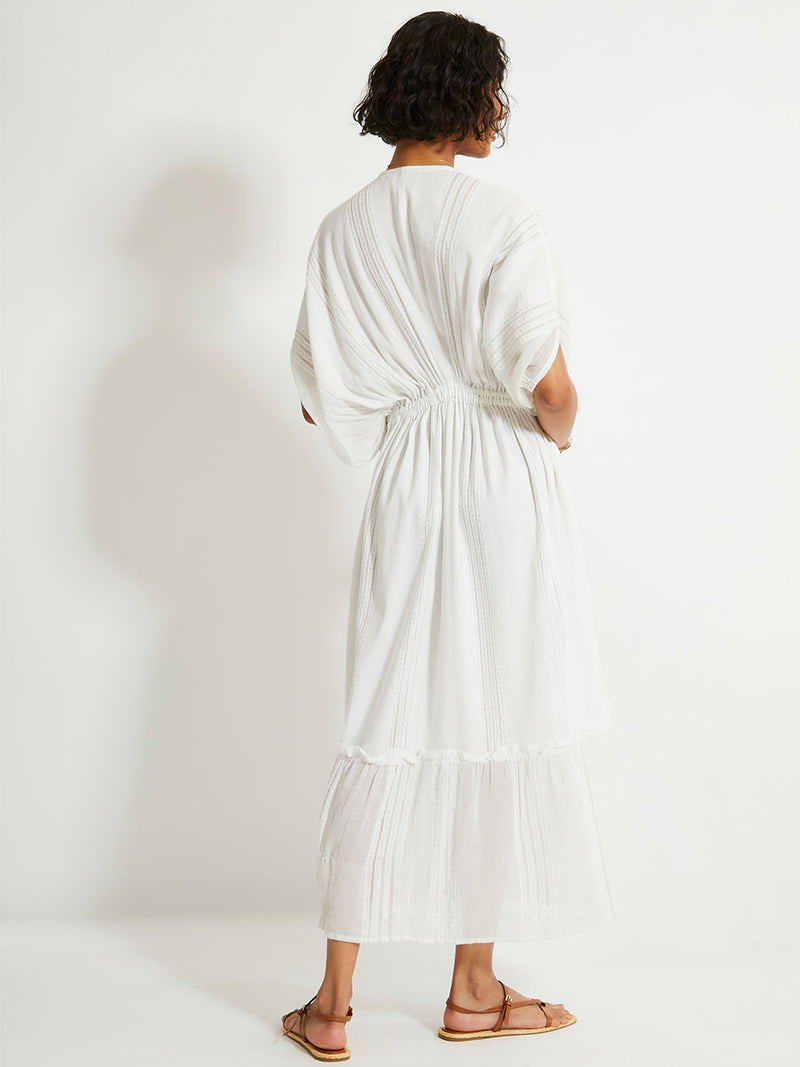 Back view of a woman standing wearing a white Abira plunge neck maxi dress with stitches of silver lurex