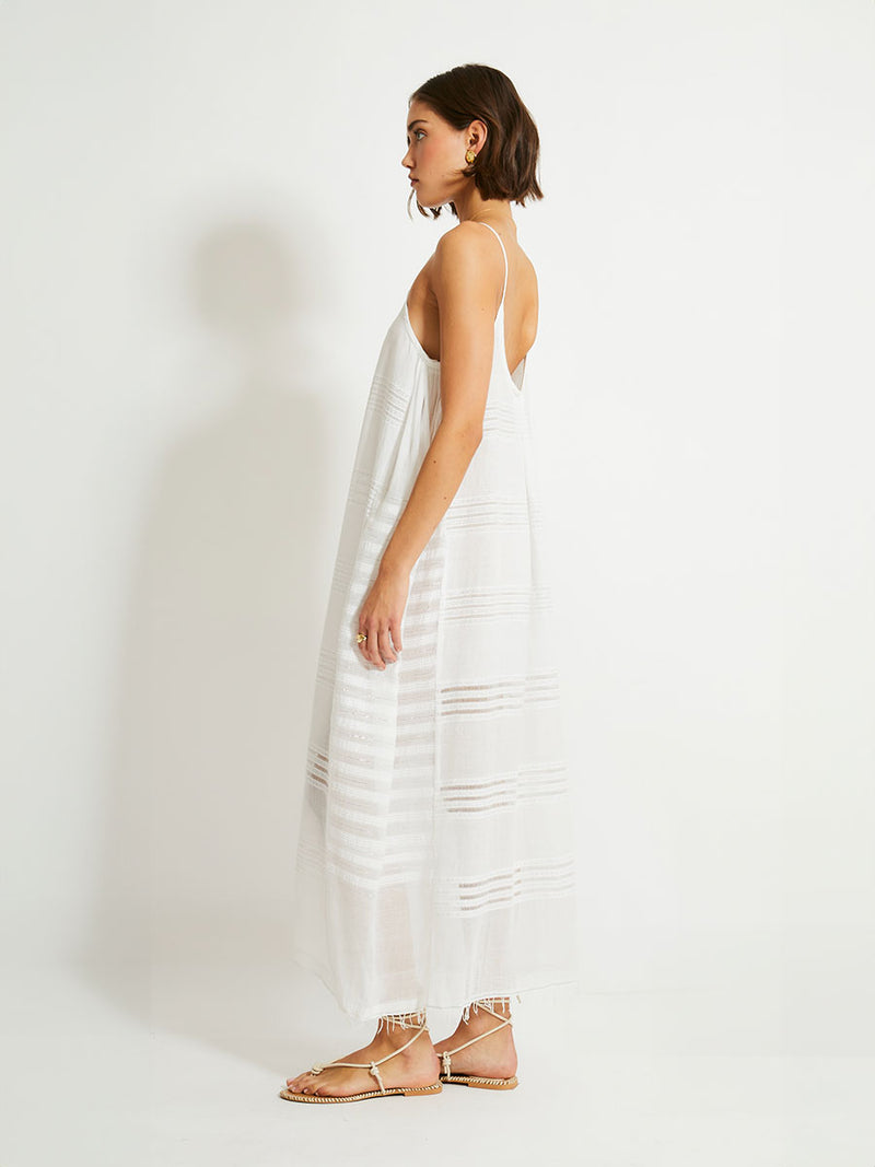 Side view of a woman walking wearing a white Abira long slip dress with spaghetti straps and stitches of silver lurex