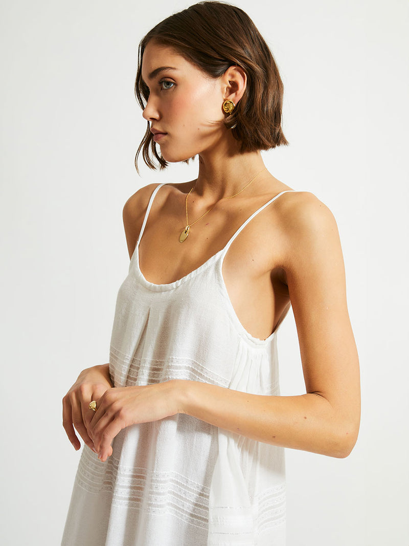 Side close up view of a woman standing with her hand in her hair wearing a white Abira long slip dress with spaghetti straps and stitches of silver lurex