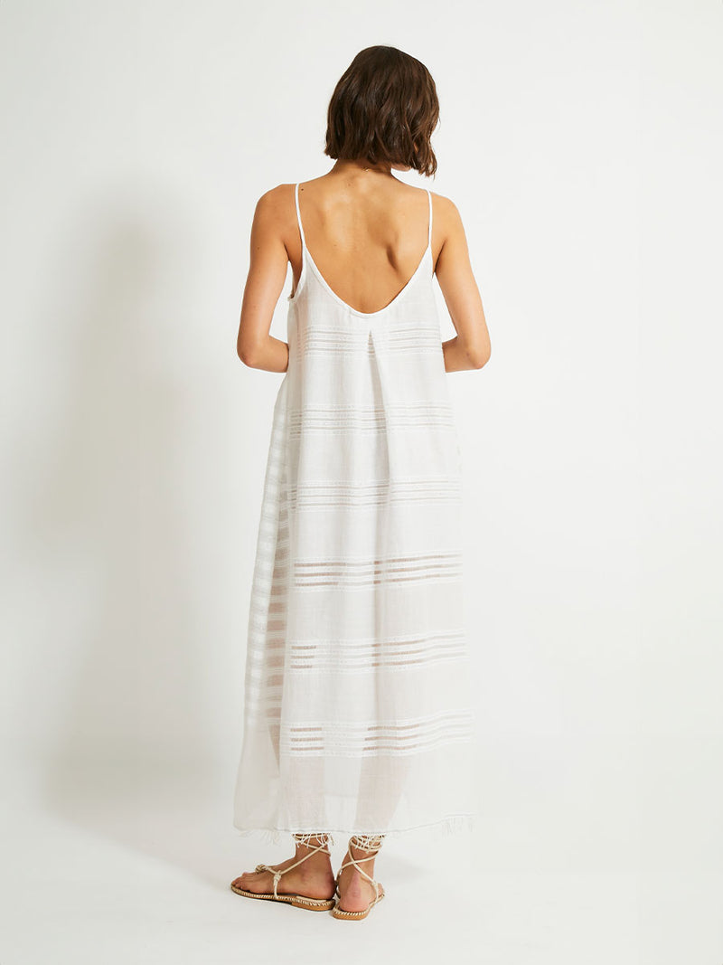 Back view of a woman standing wearing a white Abira long slip dress with spaghetti straps and stitches of silver lurex