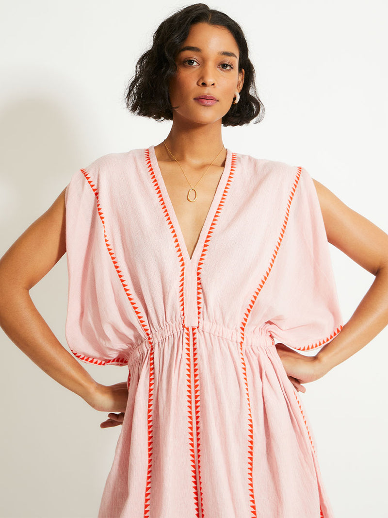 Close up on a Woman Standing Wearing lemlem ALEM Plunge Dress  featuring vibrant orange red Tibeb pattern on a soft antique rose pink background, with a hint of citron tipping.