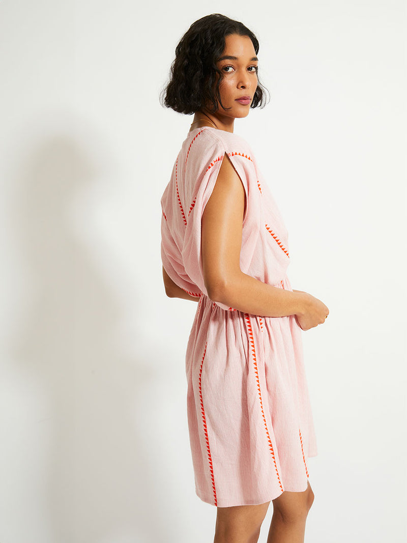 Side of a Woman Standing Wearing lemlem ALEM Plunge Dress  featuring vibrant orange red Tibeb pattern on a soft antique rose pink background, with a hint of citron tipping.