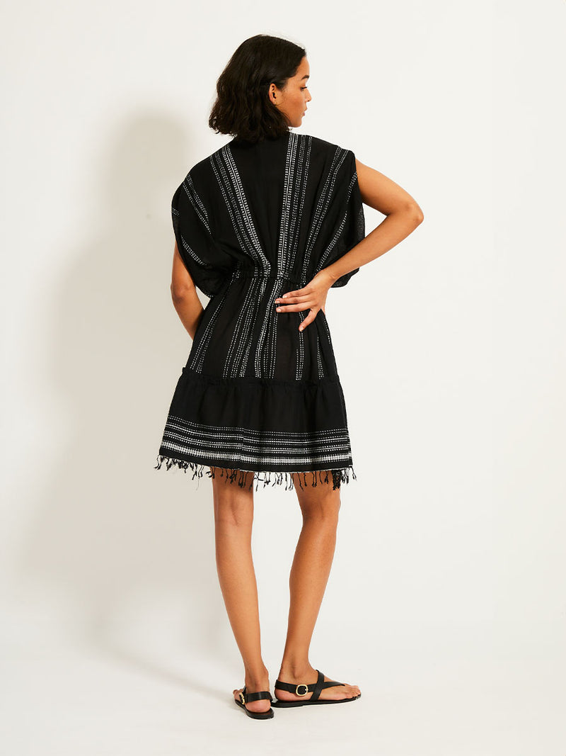 Back view of a woman standing with her hand on a white wall wearing the Leliti Short Plunge Neck Dress in Black with white stitching allover.