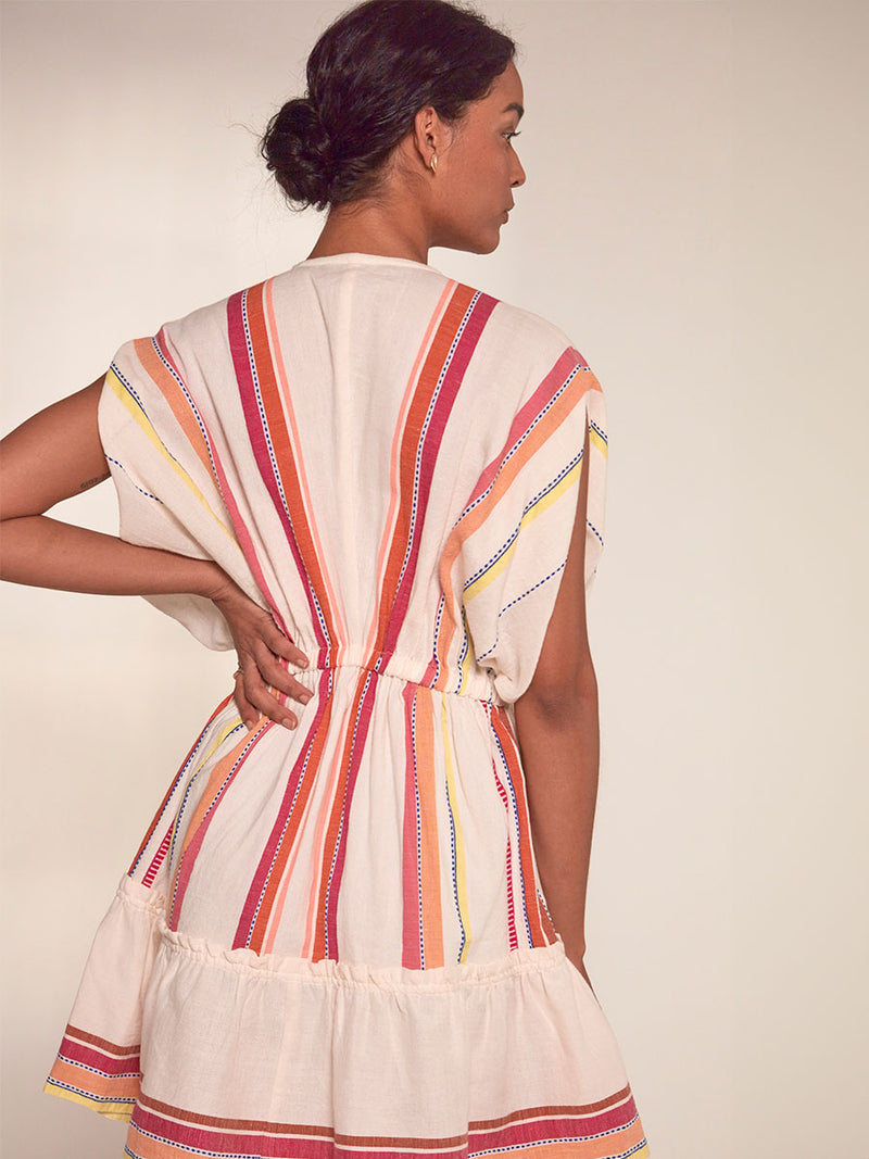 Back View of a Woman Standing Wearing lemlem Alem Plunge Dress featuring tibeb inspired stripes in a vibrant fiesta of colors against a creamy vanilla background.