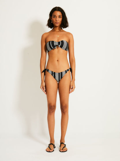 Woman standing wearing the Luchia Bandeau Top and matching Side Tie Bikini Bottom in black white white graphic diamond and arrows pattern.