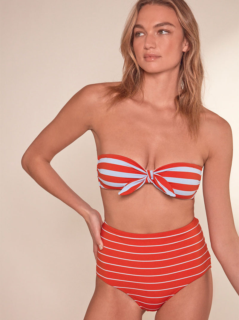 Woman Standing wearing lemlem Ava Bandeau Top featuring bold and bright tangerine color, accented by pale blue stripes and lemlem high waisy bikini bottom in jeliba tangerine color