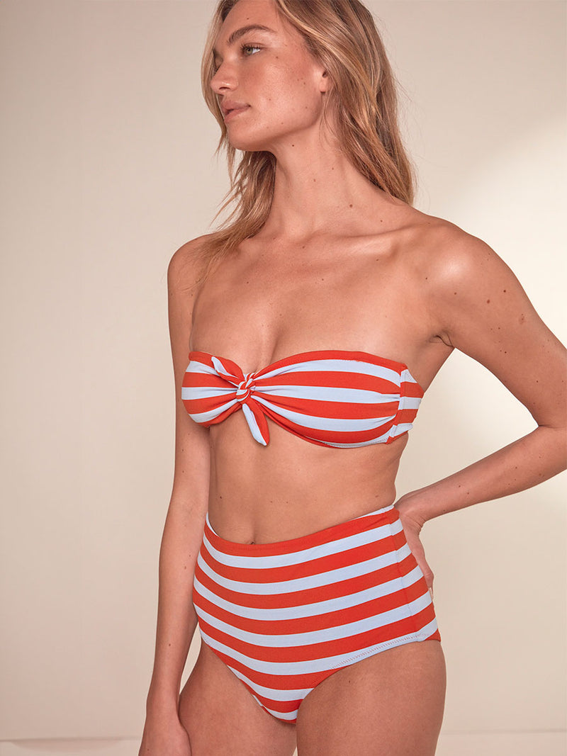 Side shot of a  Woman Standing wearing lemlem Ava Bandeau Top featuring bold and bright tangerine color, accented by pale blue stripes and a matching lemlem high waist bikini bottom