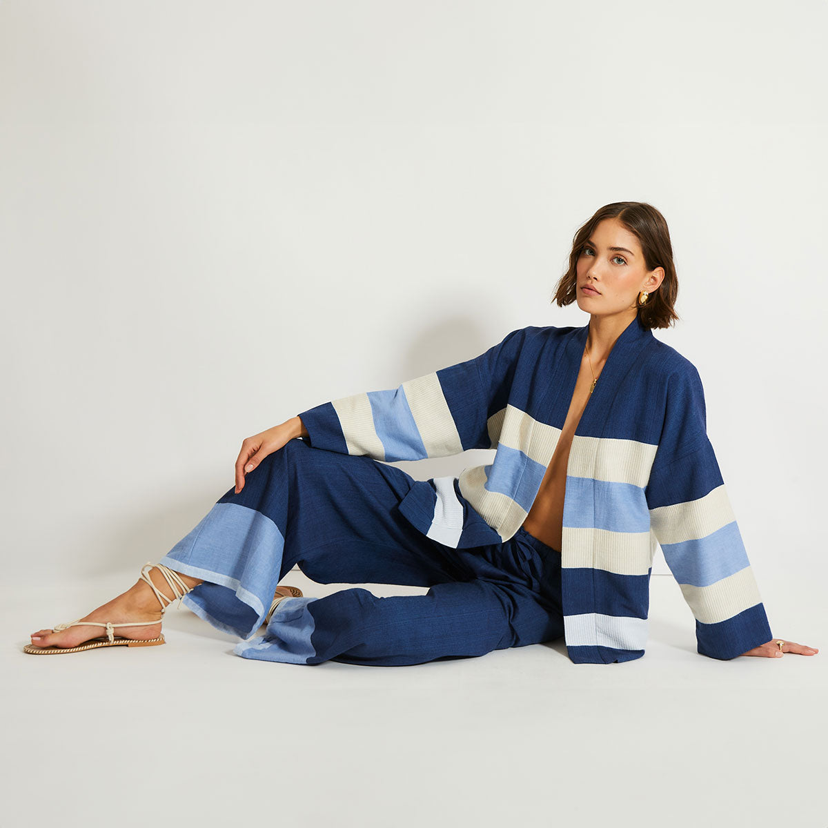 Woman sitting on a white floor wearing a blue lemlem Terara jacket with matching lemlem desta pants in navy blue. 