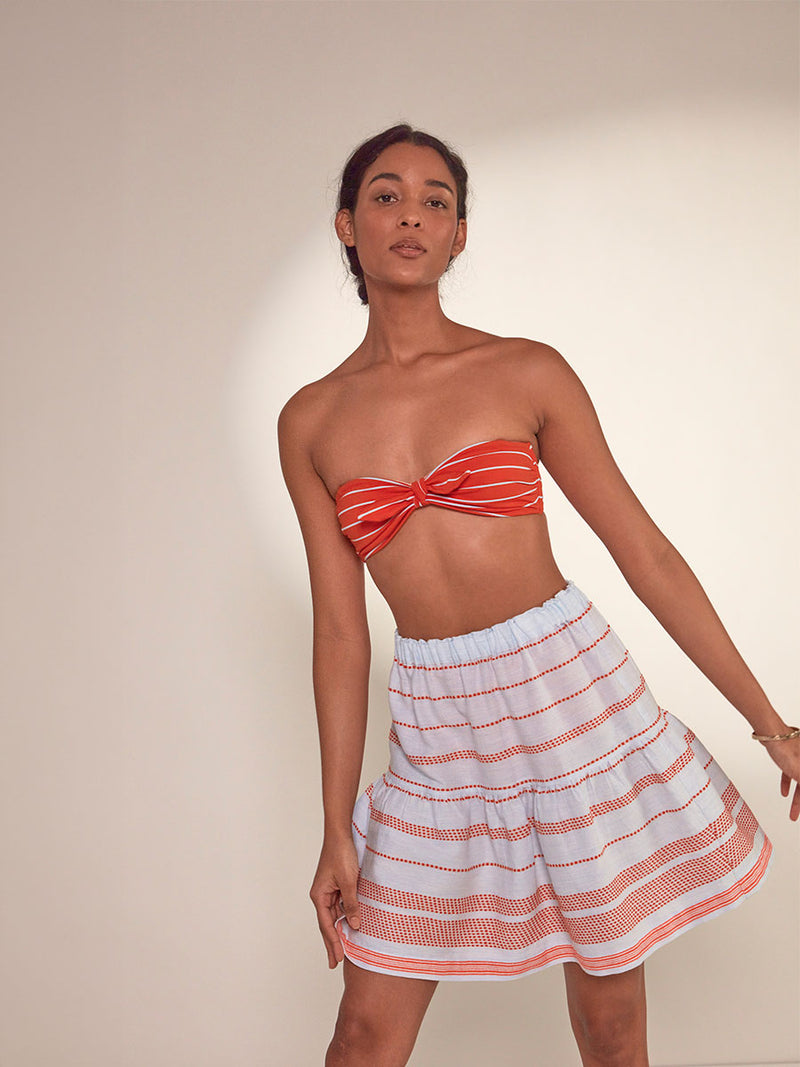Woman Standing Wearing lemlem Cheri Skirt featuring playful pattern of red dots becoming stripes on a pale blue background and Jeliba Tangerine Ava Bandeau Top