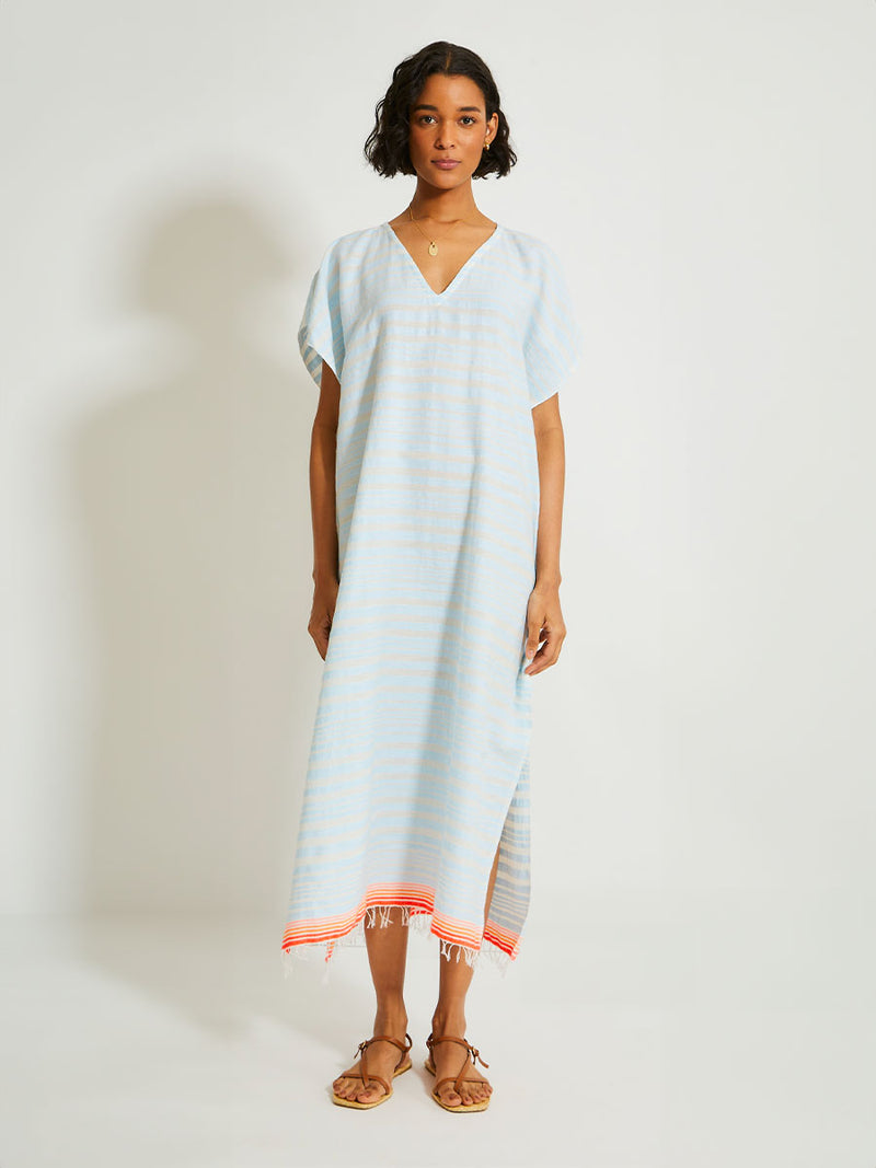 Woman Standing Wearing lemlem DALILA Caftan featuring a soft, variegated two-color stripe in sky blue and cream, complemented by placed bands of bright orange and tangerine