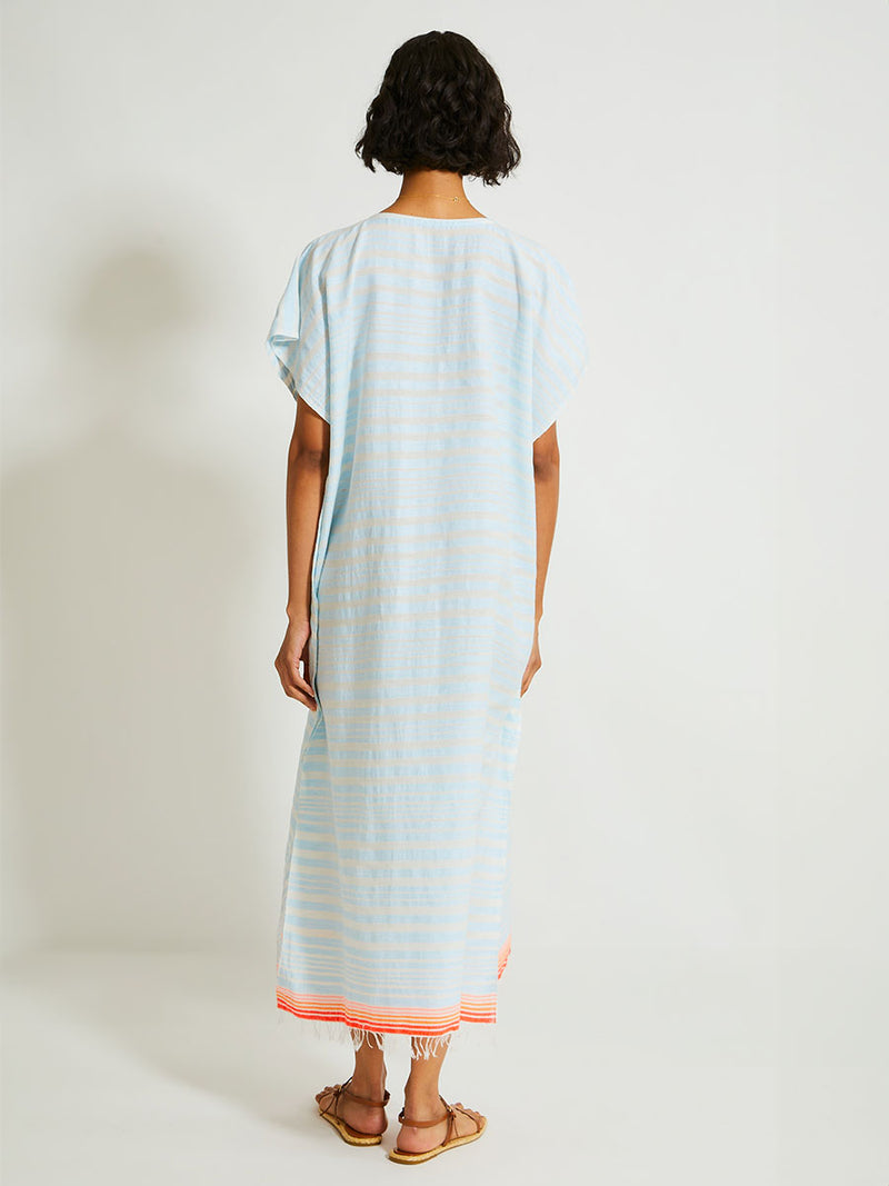 Back of a Woman Standing Wearing lemlem DALILA Caftan featuring a soft, variegated two-color stripe in sky blue and cream, complemented by placed bands of bright orange and tangerine