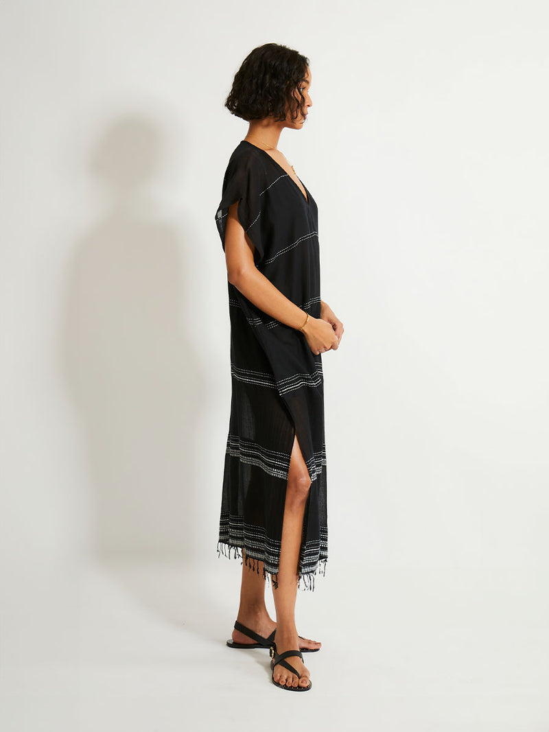 Side view of a woman standing wearing the Leliti Classic Caftan in Black with white stitching allover.