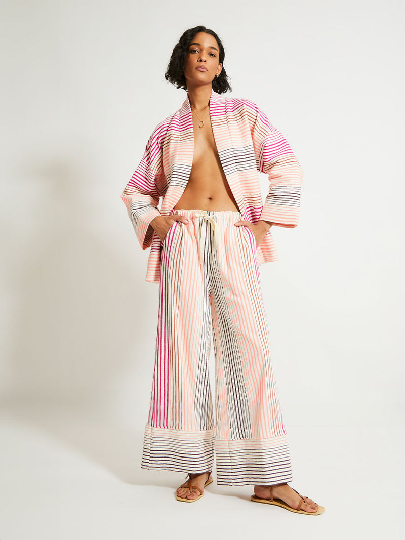 Woman Standing Wearing lemlem Terarra Jacket and matching Wide Leg Pants featuring ombre stripe pattern, where deep plum seamlessly transitions into peach, chestnut, and bright pink