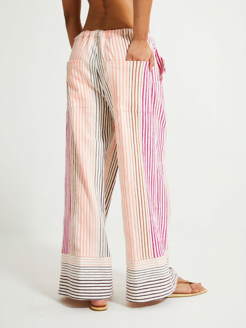 Back of a Woman Standing Wearing lemlem Wide Leg Pants featuring ombre stripe pattern, where deep plum seamlessly transitions into peach, chestnut, and bright pink