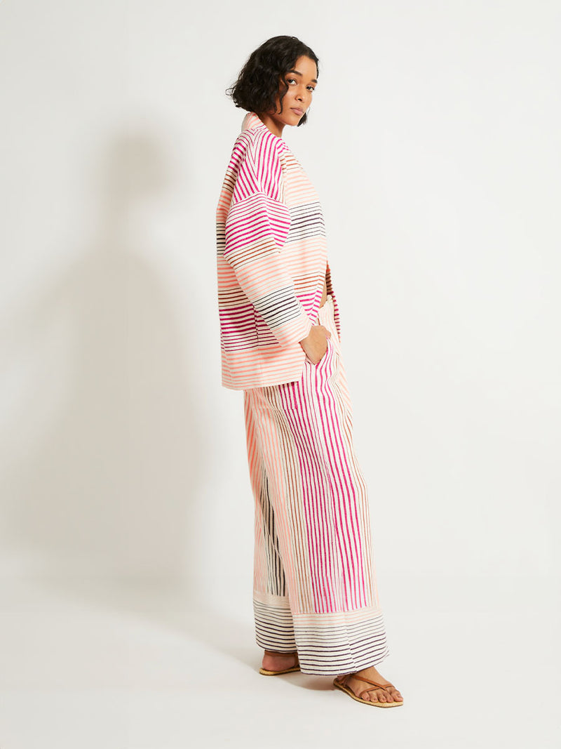 Side view on a Woman Standing Wearing lemlem Terarra Jacket and matching Wide Leg Pants featuring ombre stripe pattern, where deep plum seamlessly transitions into peach, chestnut, and bright pink